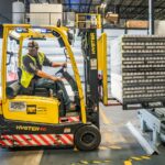 Magazijnmedewerker verplaatst stapel blikjes op kunststof pallet met heftruck in logistieke hal