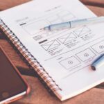Schets van een websiteontwerp in een notitieboek, naast een pen en smartphone op een houten tafel